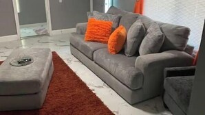 Ein Wohnzimmer mit grauem Sofa, orangefarbenen Kissen, einem roten Teppich und einem Marmorboden.