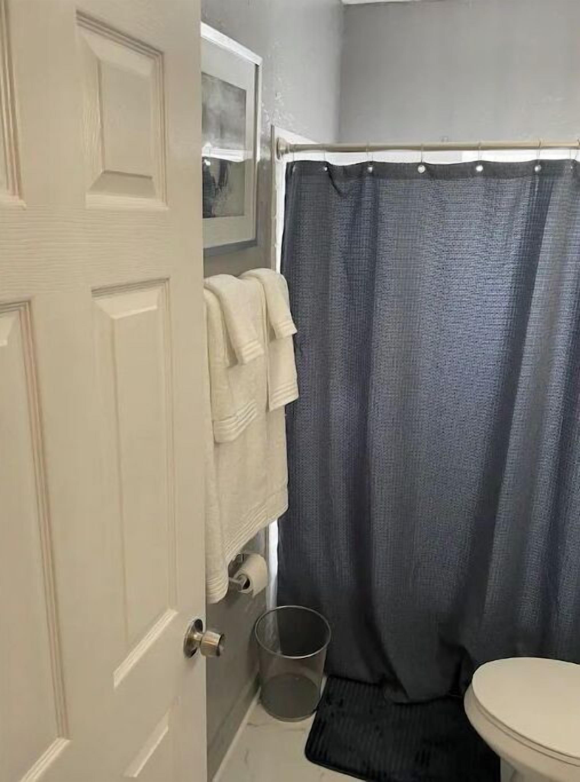 A bathroom with a white door, a shower curtain, a towel rack with towels, a toilet, and a trash can.