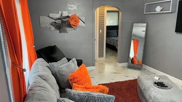 Ein Wohnzimmer mit grauem Sofa, orangefarbenen Kissen, einem roten Teppich und einem grauen Hocker.