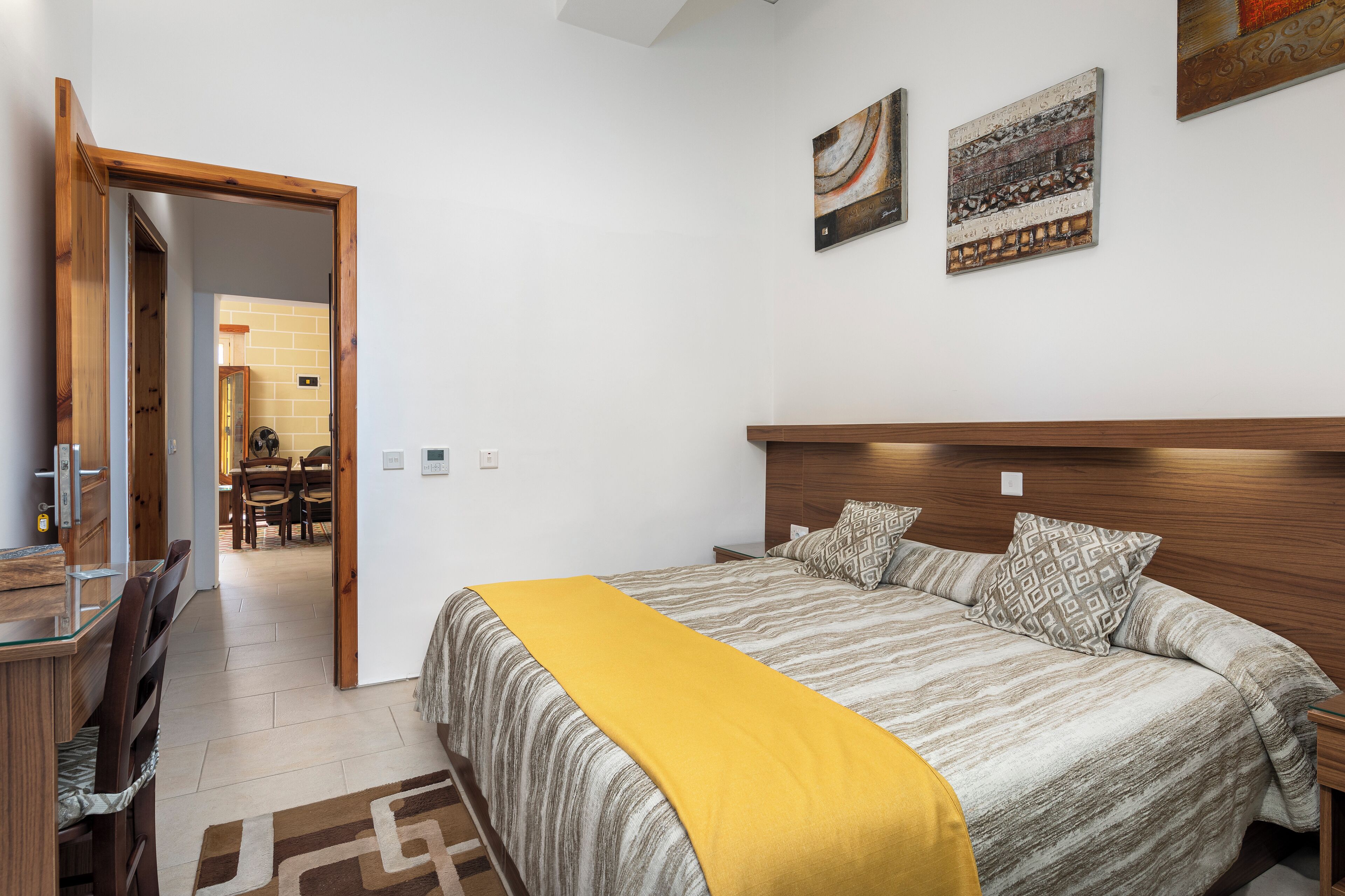 Un dormitorio con cama, cabecera de madera y una manta amarilla. Hay dos cuadros en la pared y una zona de comedor al fondo.