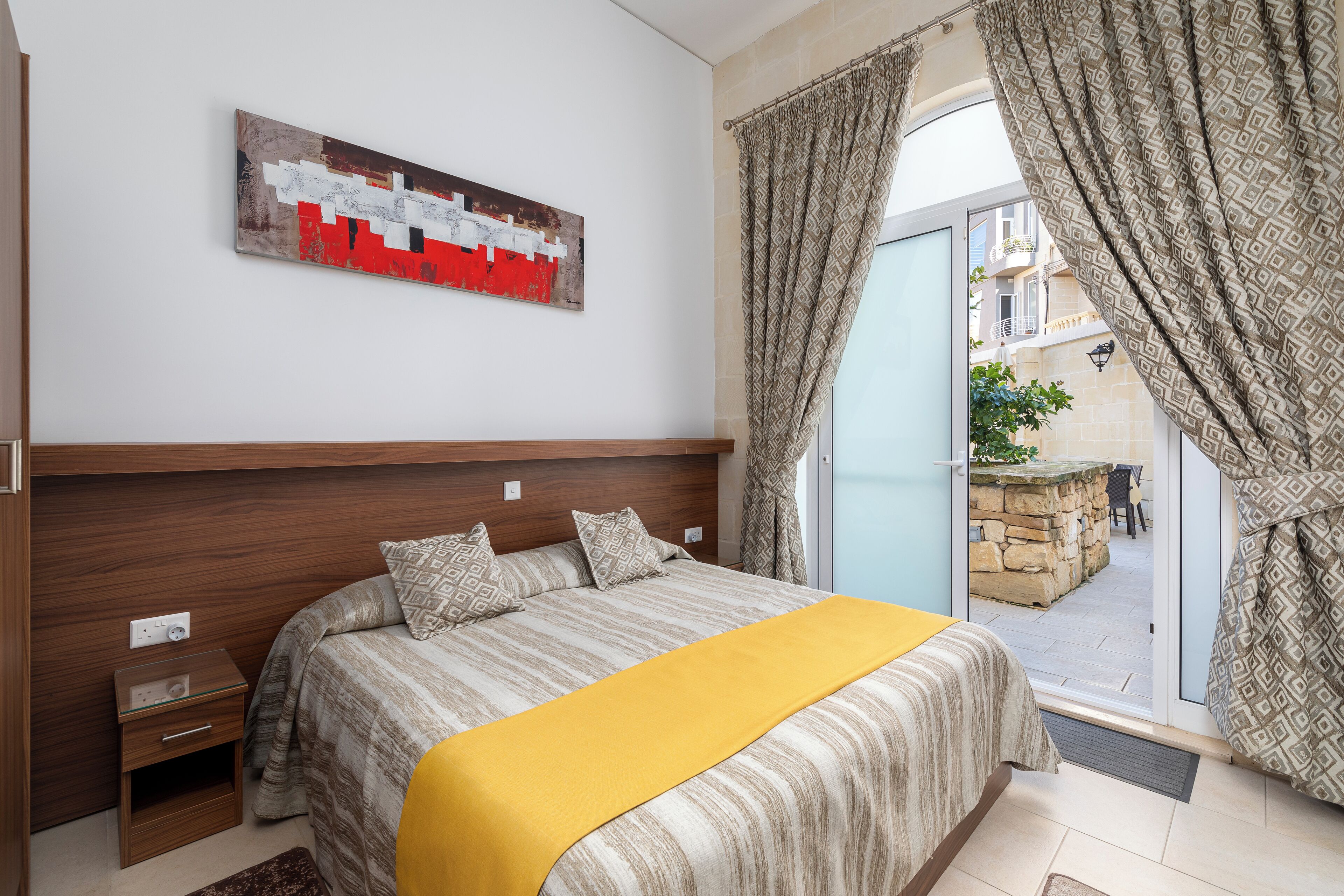 A bedroom with a bed, a bedside table, a power outlet, a window with curtains, and a view of the outside.