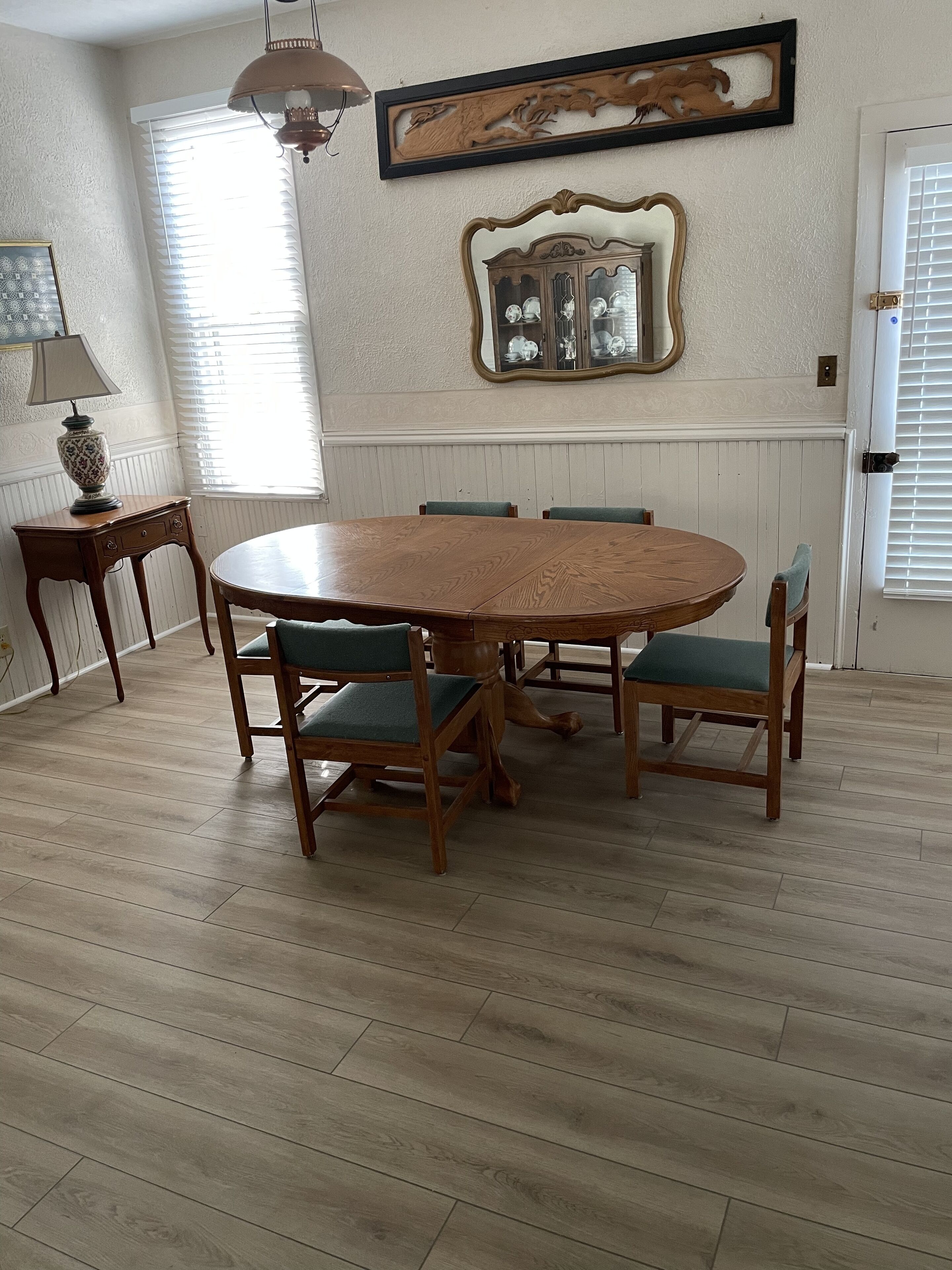 Une salle à manger avec une table ronde en bois entourée de six chaises dotées de coussins verts, une table d’appoint en bois avec une lampe, et un miroir décoratif accroché au mur.