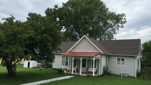 Une maison de plain-pied avec un toit rouge, une véranda aux colonnes blanches et un grand arbre devant.