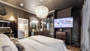 Ein modernes Schlafzimmer mit einem großen Bett, einem Fernseher, einer Chandelüster und einem durch eine offene Tür einsehbaren Badezimmer.