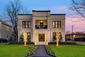 Une maison moderne à deux étages, dotée d’une entrée centrale, d’un balcon à l’étage supérieur et d’une pelouse bien entretenue agrémentée d’un éclairage décoratif.