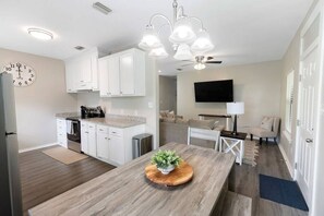 A modern kitchen with white cabinets, a wooden dining table, and a ceiling fan.