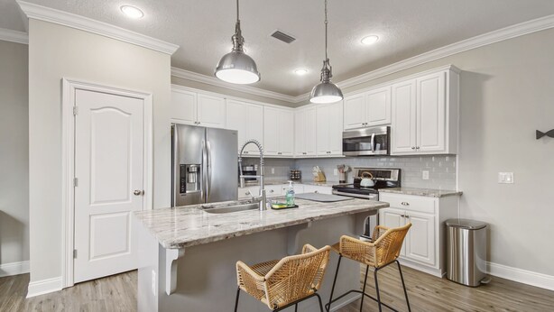A kitchen with white cabinetry, stainless steel appliances, a marble countertop, and two wicker bar stools. - Cozy home in a great location. Pool, ping-pong and arcade! (Panama City Beach)