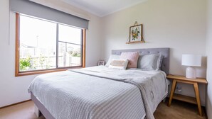 Un dormitorio con un ventanal grande, una cama con cabecera gris, una mesita de noche con lámpara y un cuadro enmarcado en la pared.