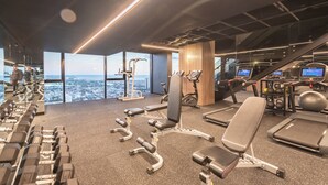 Ein modernes Fitnessstudio mit verschiedenen Trainingsgeräten, einem großen Fenster mit Stadtblick und einer Treppe, die in eine obere Ebene führt.