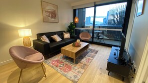 Ein modernes Wohnzimmer mit einem schwarzen Sofa, zwei rosafarbenen Sesseln, einem Holztisch und einem großen Fenster mit Blick auf die Stadt.