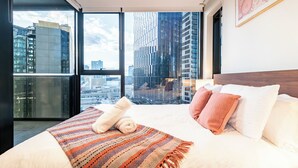 Ein modernes Hotelzimmer mit einem großen Bett und Blick auf die Stadt durch raumhohe Fenster. Im Hintergrund ist der Stadthimmel zu sehen.