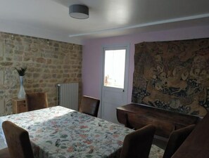 A dining area with a table covered in a floral tablecloth, surrounded by wooden chairs. A stone wall and a door with a window are visible in the background.