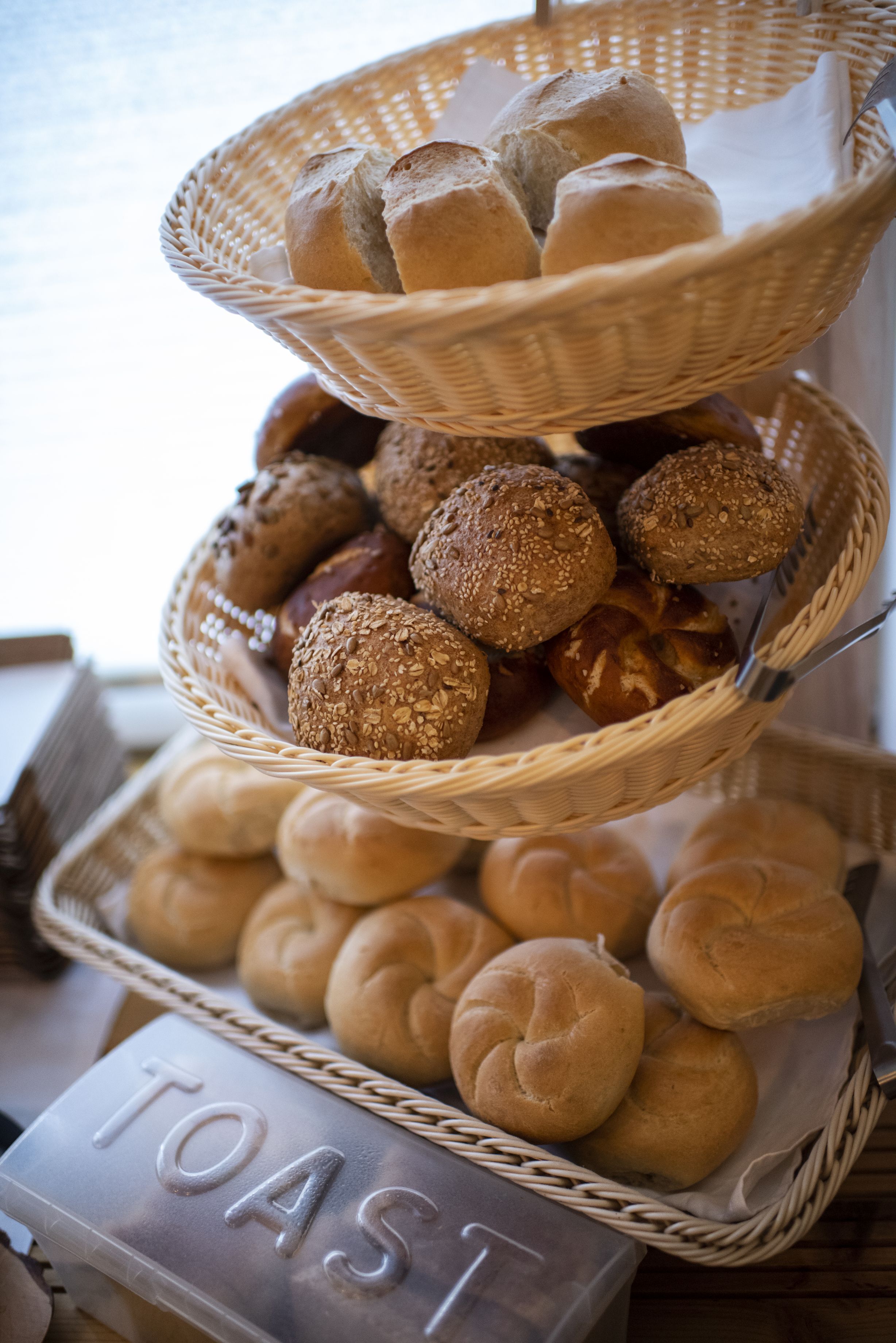 En korg i flera våningar, fylld med olika sorters bröd, inklusive frallor och bullar, placerad på ett bord med en skylt som visar texten "ROSTAT BRÖD".