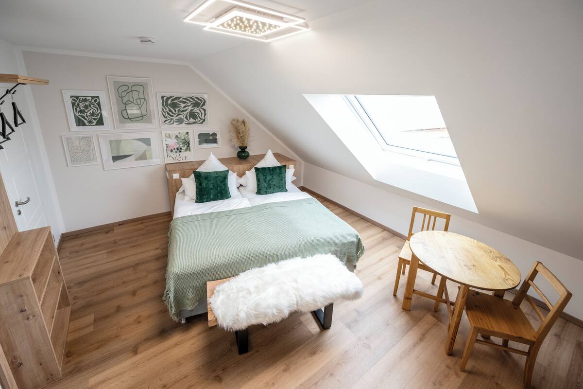 Ein Schlafzimmer mit einem Bett, einer Bank, einem runden Tisch und Stühlen. Es gibt ein Oberlicht und Kunstwerke an der Wand.