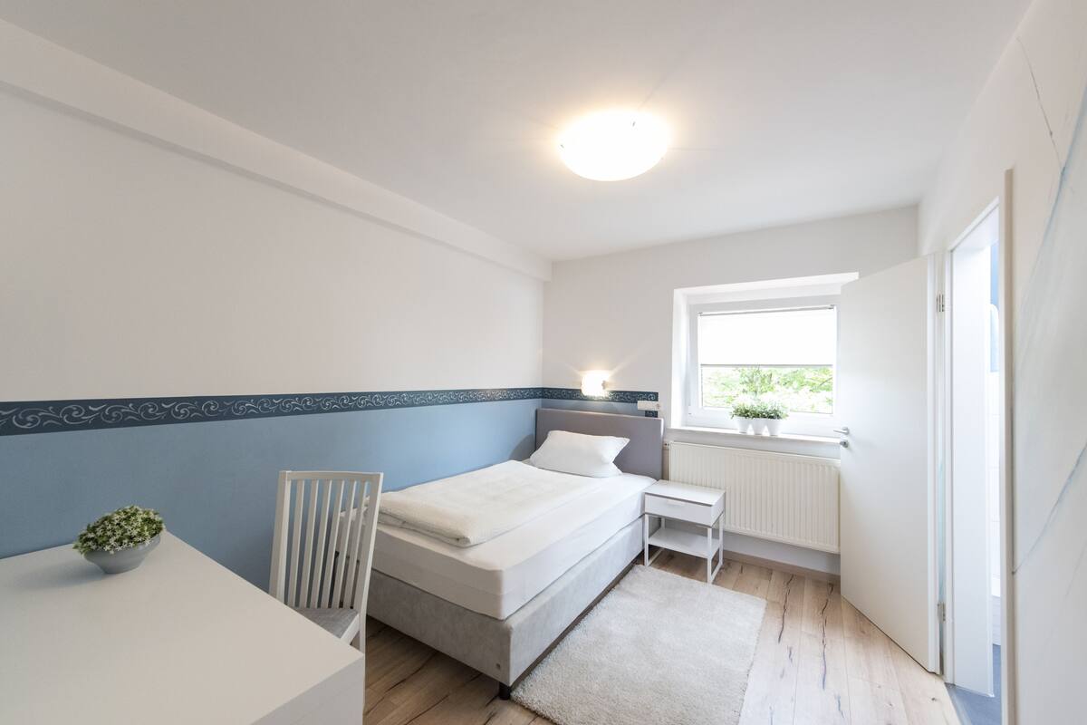 Ein Schlafzimmer mit Bett, Nachttisch, Stuhl und einem Fenster mit Blick auf Grünflächen.
