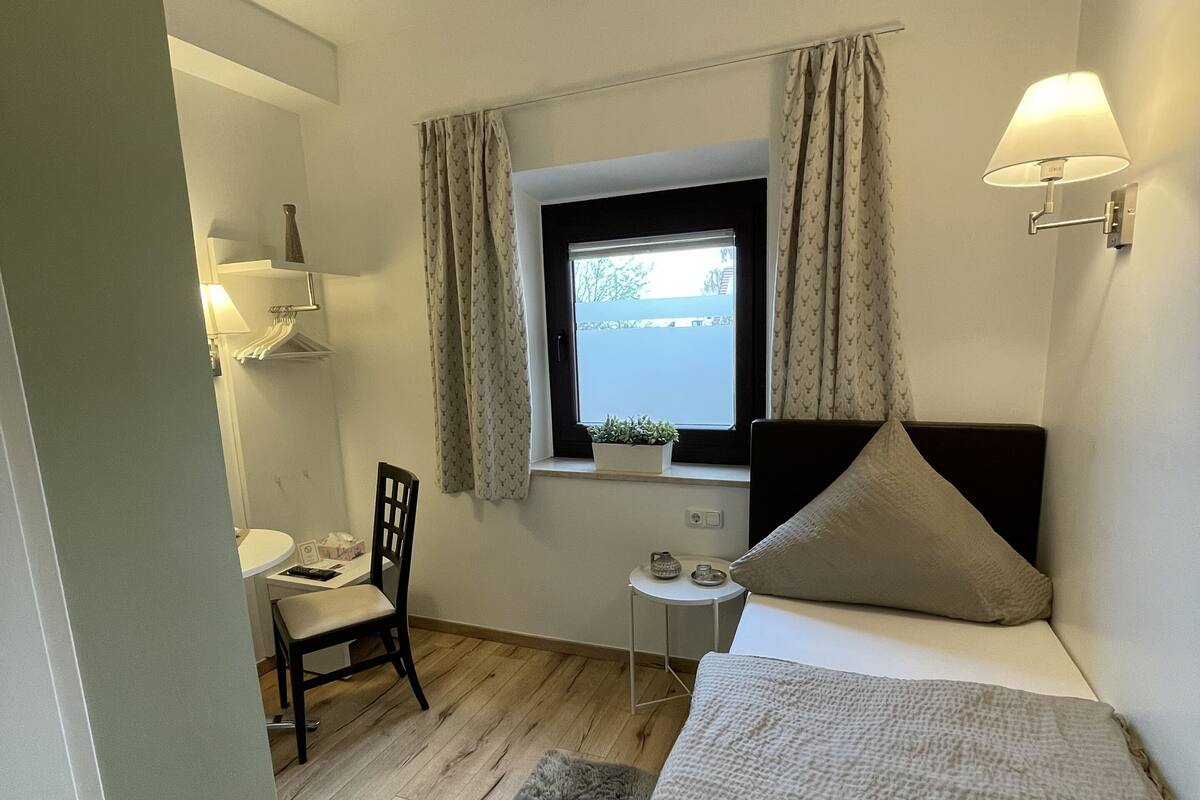 Ein gemütliches Schlafzimmer mit einem Bett, einem kleinen Schreibtisch, einem Sessel, einem Fenster mit Vorhängen und einer Wandlampe.