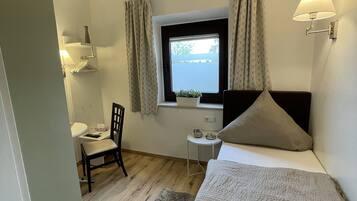 Ein gemütliches Schlafzimmer mit einem Bett, einem kleinen Schreibtisch, einem Sessel, einem Fenster mit Vorhängen und einer Wandlampe.