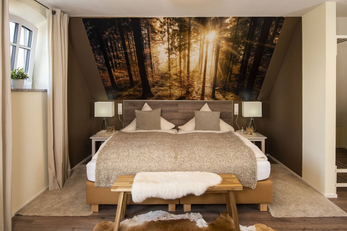 Ein Schlafzimmer mit einer großen Waldlandschaftsmalerei, einem Bett mit Holz-Kopfteil, Nachttischen mit Lampen und einer kleinen Bank.