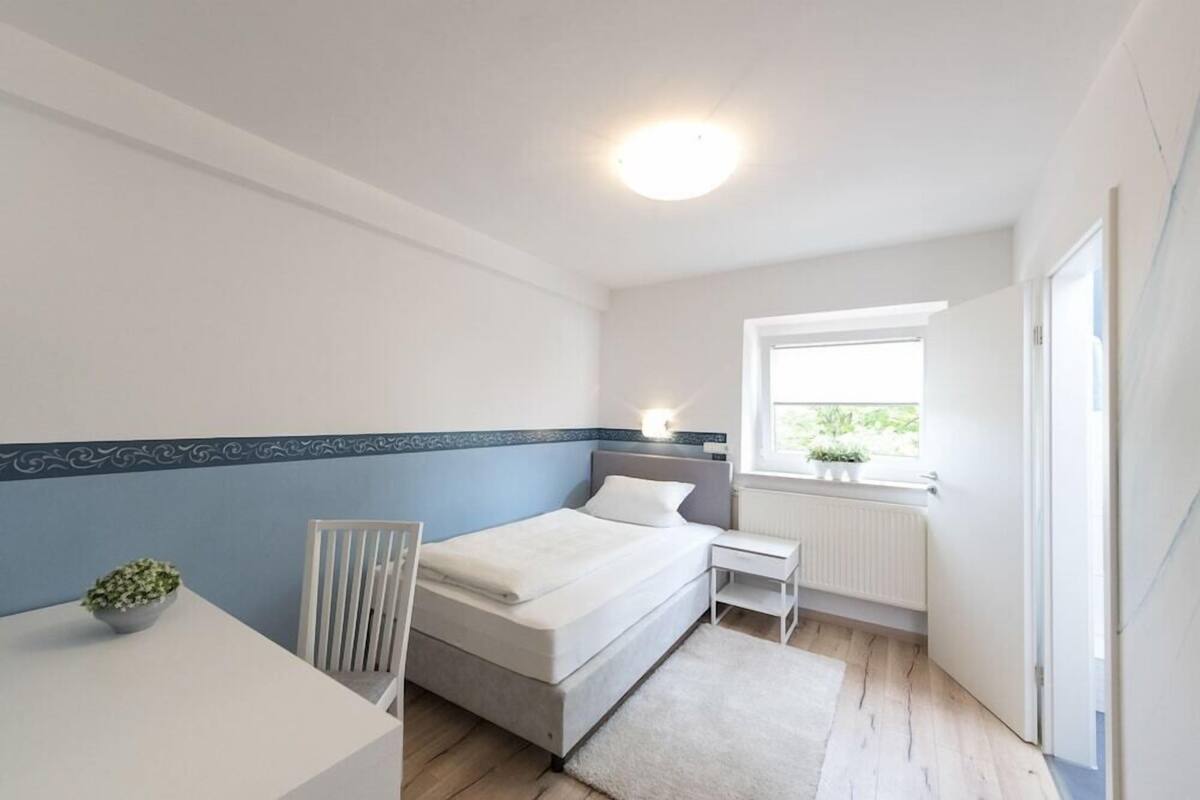 Ein Schlafzimmer mit Bett, Nachttisch, Stuhl, Fenster, Heizkörper und einem kleinen Tisch mit Pflanze.