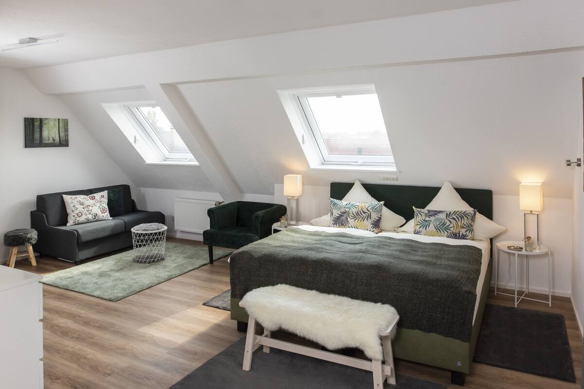 Ein modernes Schlafzimmer mit einer Loft-Decke, einem großen Bett mit vielen Kissen, einer Bank, einem kleinen Sofa und zwei Nachttischen mit Lampen.