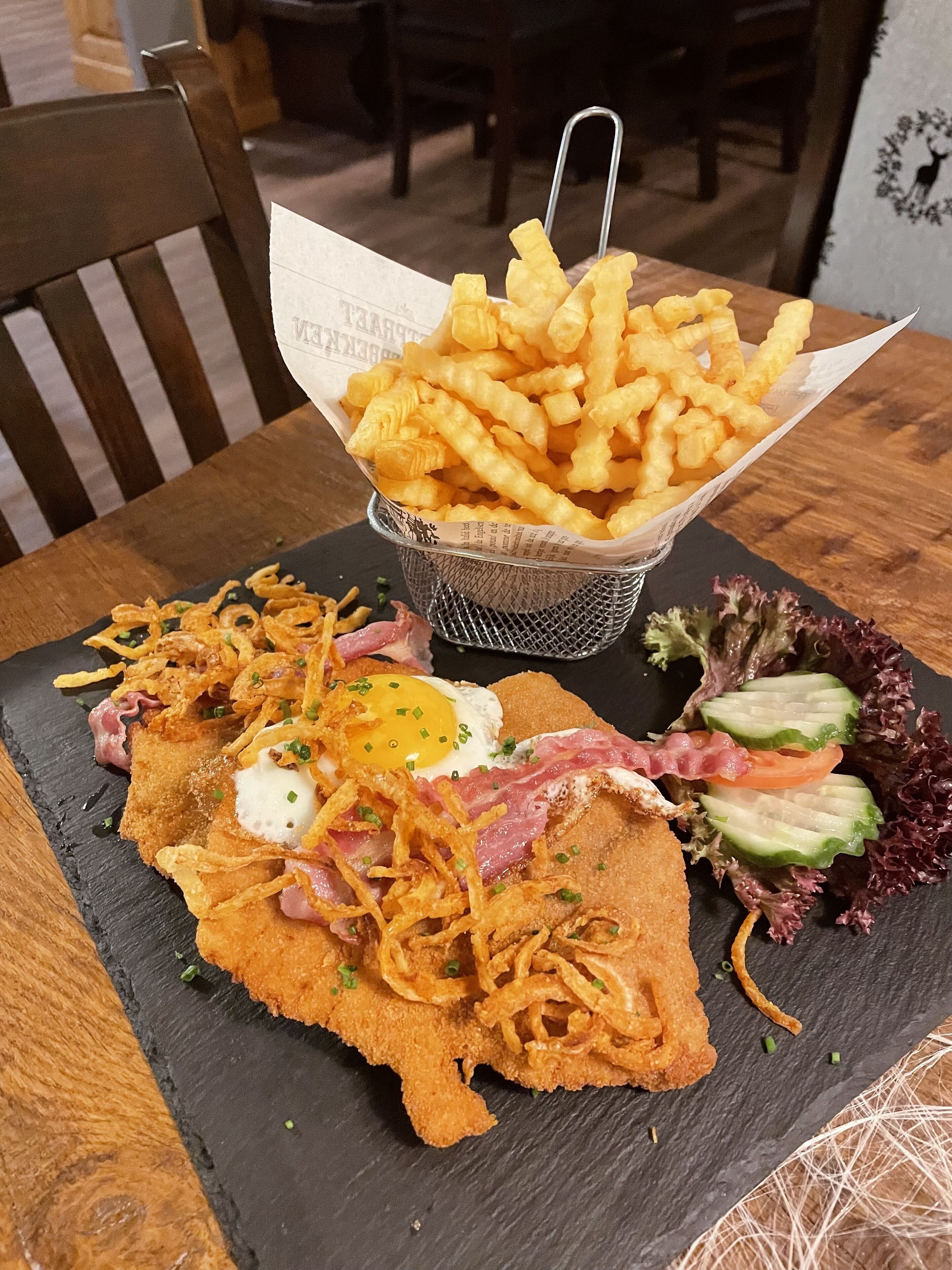 A meal consisting of a fried egg, bacon, and a salad on a slate plate, with a basket of fries in the background.