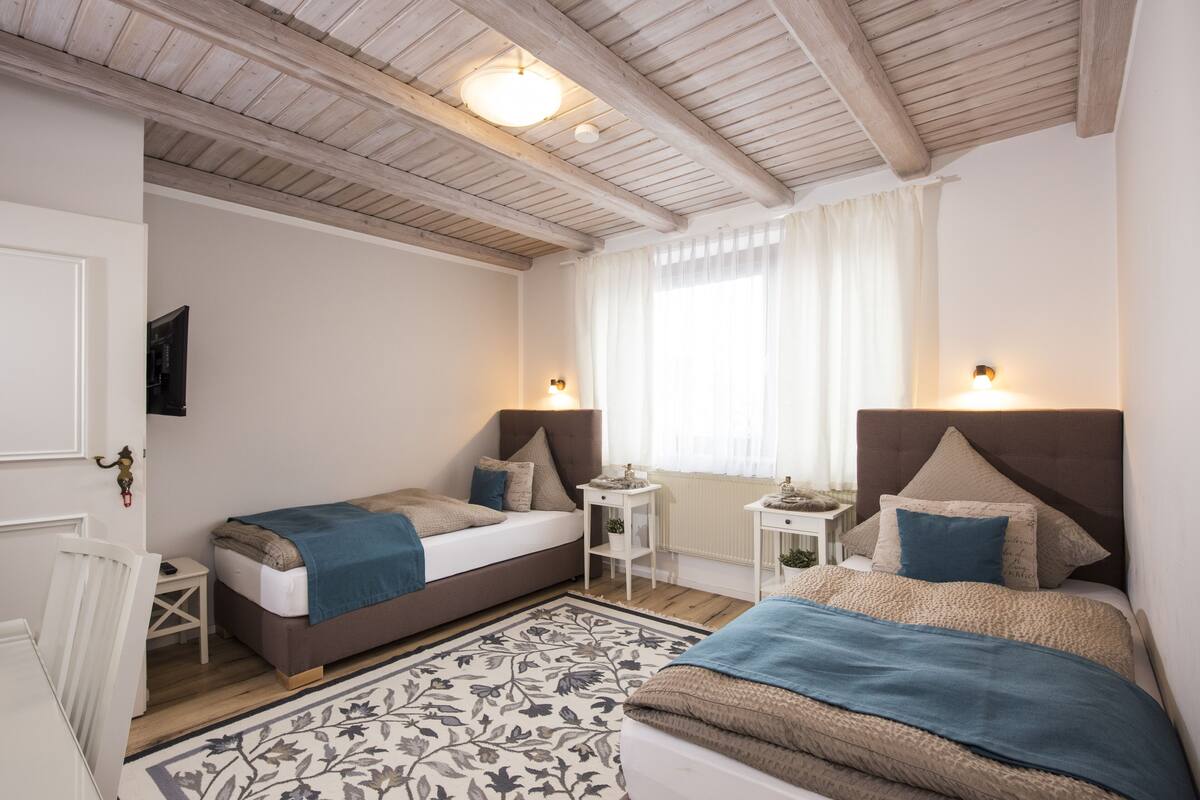 Ein Schlafzimmer mit zwei Betten, einem kleinen Nachttisch, einem Beistelltisch, einem gemusterten Teppich und einer Decke mit freiliegenden Holzbalken.