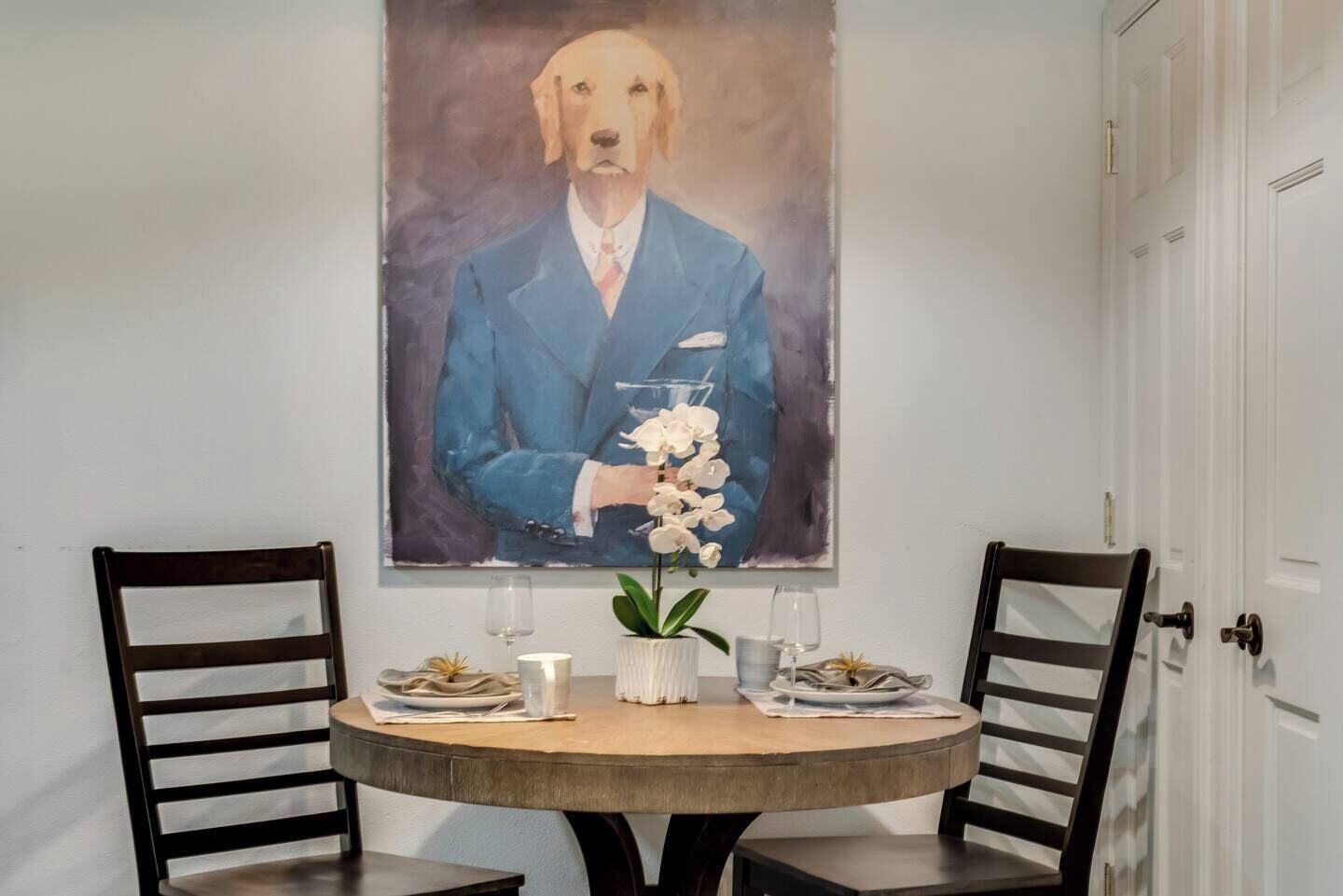 Un comedor con una mesa redonda, dos sillas, un cuadro de un perro con traje y un jarrón con flores.