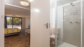 Baño con ducha, inodoro y puerta de cristal que da a un dormitorio con cama y televisor.