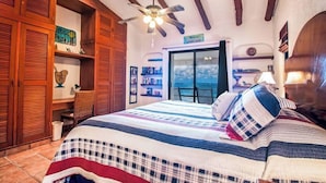 Une chambre à coucher avec un lit, une armoire en bois, un ventilateur de plafond et une vue sur la mer, visible par une porte.