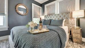 A bedroom with a tufted headboard, a bedside table with a lamp, and a tray with a bottle, flowers, and glasses on the bed.