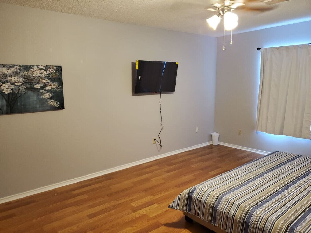 A bedroom with hardwood flooring, a bed with a striped bedspread, a ceiling fan, a mounted flat-screen TV, and a painting on the wall.