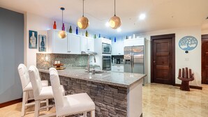 Una cucina moderna con mobili bianchi, un piano di lavoro in granito e una parete decorata in pietra. Si notano tre lampade a sospensione, un frigorifero e un forno a microonde.