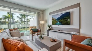 A modern living room with a large TV, a sofa, a coffee table, and a view of a pool and greenery outside. - New modern villa, with pool, 5 minutes from Disney (Kissimmee)