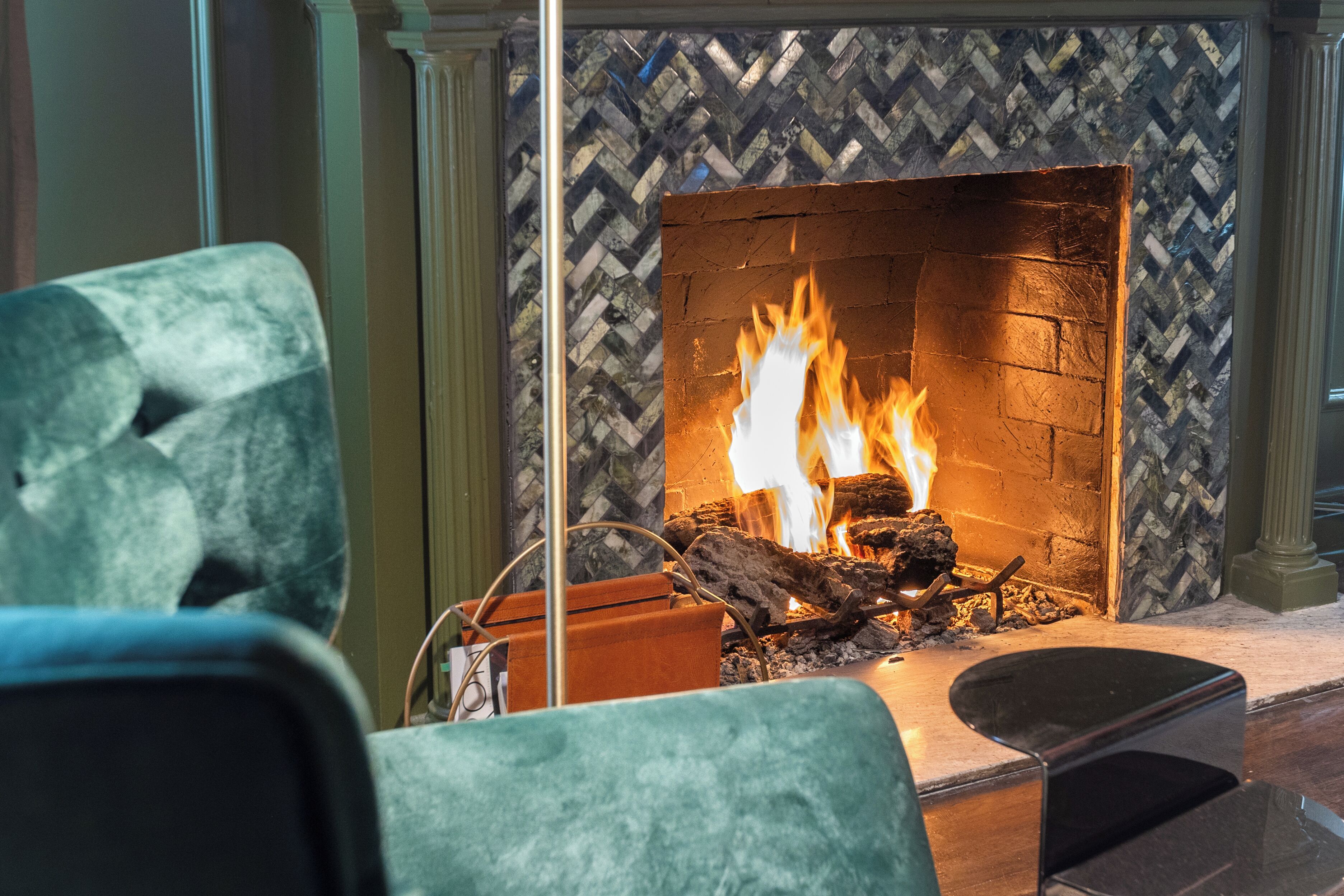 A fireplace with a herringbone-patterned wall, a fire burning, and a seating area with a blue chair in the foreground.