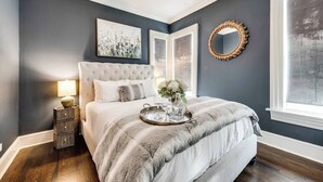 Una camera da letto con una testiera trapuntata, un comodino con una lampada, un'ampia finestra e uno specchio decorativo.