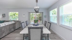 Un coin repas avec une table blanche, des chaises et un lustre. On aperçoit un canapé avec des coussins à l’arrière-plan.