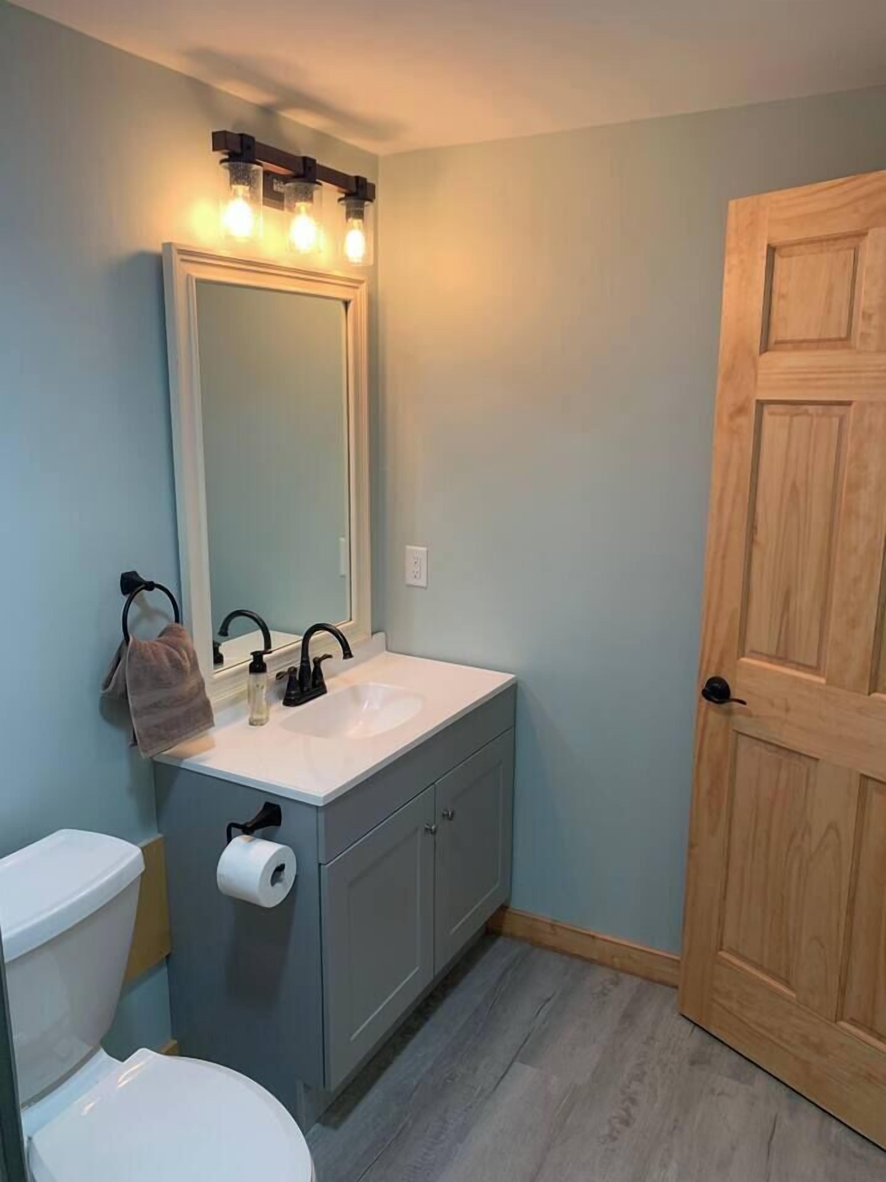 Un baño con lavamanos blanco, un espejo con iluminación, un perchero para toallas y una puerta de madera.