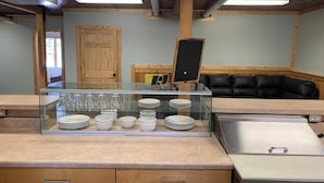 A kitchen area with a glass display case containing dishes and glasses, wooden cabinets, and a black leather couch in the background.