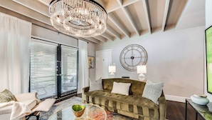 A living room with a chandelier, a round clock, a sofa, and a glass coffee table. - Spacious 1-bed executive condo on Dallas’ historic Swiss Avenue. Enjoy the porch access, high-speed Wi-Fi, king bed with Egyptian-cotton linens, and modern design with upscale finishes—perfect for morning coffee, remote work, or relaxing evening (Dallas)