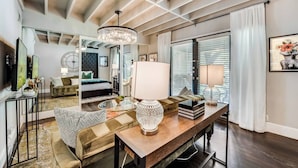 A modern living room with a large chandelier, a sofa, a dining table, and a view of the bedroom through a glass door. - Spacious 1-bed executive condo on Dallas’ historic Swiss Avenue. Enjoy the porch access, high-speed Wi-Fi, king bed with Egyptian-cotton linens, and modern design with upscale finishes—perfect for morning coffee, remote work, or relaxing evening (Dallas)