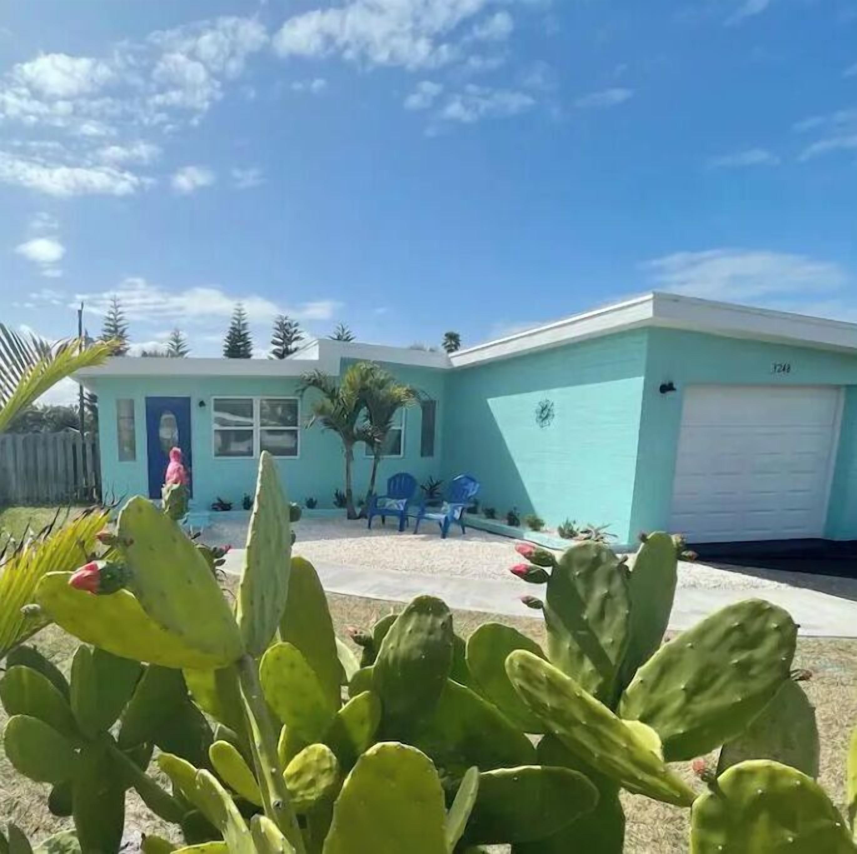 Une maison moderne à deux étages, à la façade bleu clair, avec un garage et un petit jardin devant, agrémenté de cactus et d’un espace patio.
