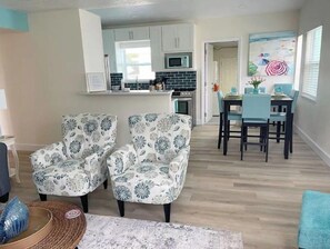 A modern living room with two patterned armchairs, a dining area with a table and chairs, a kitchen with white cabinets and a microwave, and a beach-themed painting on the wall.