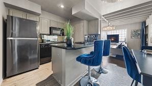 A modern kitchen with a stainless steel refrigerator, a central island with bar stools, and a dining area with a dark table and blue chairs. - Spacious 1BR/1BA condo in Dallas’ Historic Munger Place with a private fenced yard, king luxury hybrid mattress, Egyptian-cotton linens, fireplace, Smart TV, high-speed Wi-Fi, Alexa concierge, and a fully equipped gourmet kitchen. (Dallas)