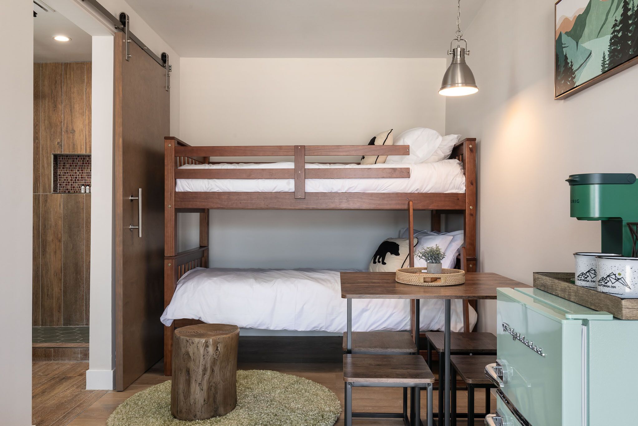 A bunk bed with a desk and chair, a wooden stool, a green appliance, and a painting on the wall.