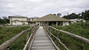 A wooden bridge leading to a building with a thatched roof, surrounded by greenery. - Hôtel Belle Terre Resort (Macouria)