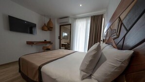 A modern hotel room with a large bed, a flat-screen TV mounted on the wall, a wooden headboard, and a small seating area with a chair and table. - Hôtel Belle Terre Resort (Macouria)