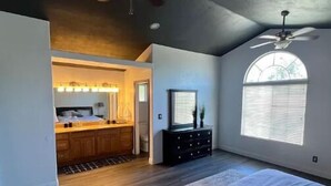 A bedroom with a large bed, a ceiling fan, a window with blinds, and a view of a kitchen area. - Newley remodeled and furnished home! (fresno)