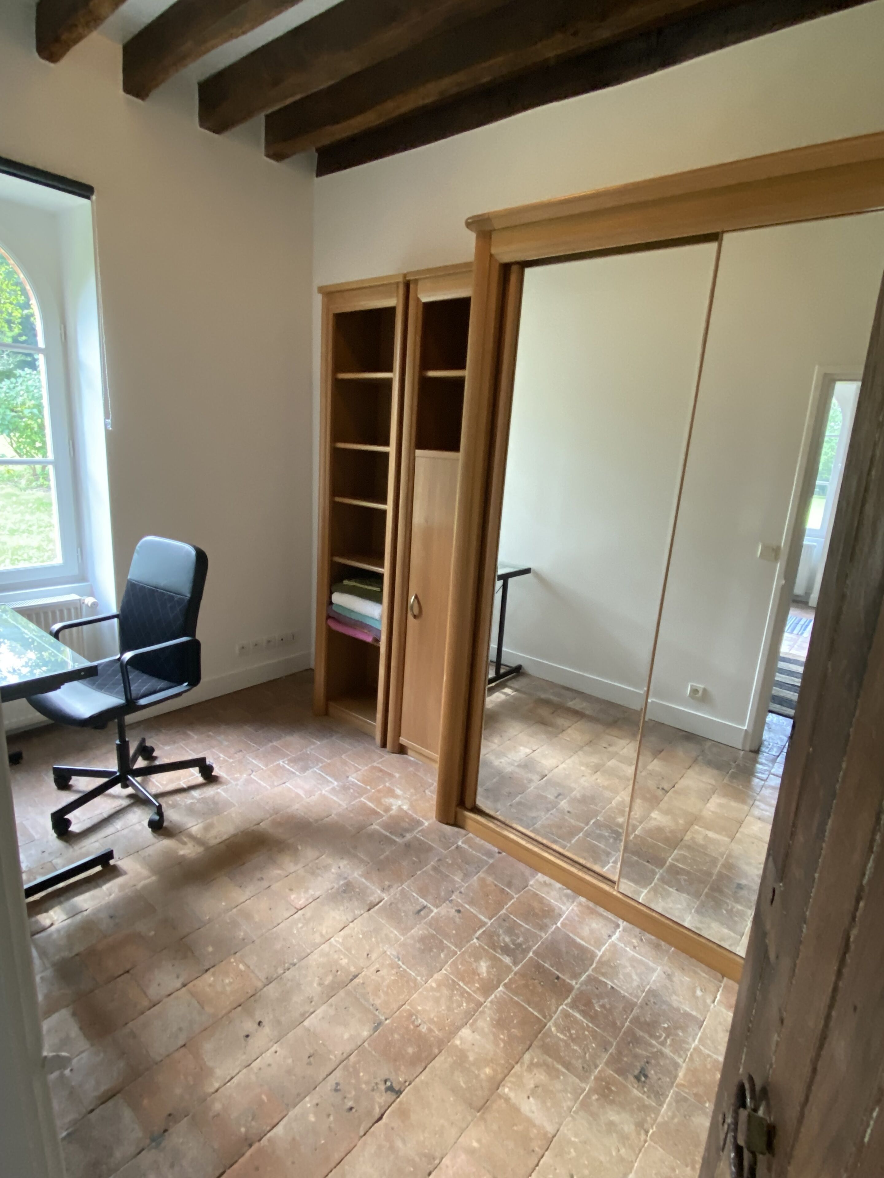 A room with a wooden floor, a desk with a chair, a wardrobe with a mirror, and a window.