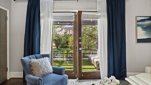 Et værelse med en blå lænestol, en hvid sofa, en glasdør, der fører til et udendørs område med et bord og stole, samt et maleri på væggen.
