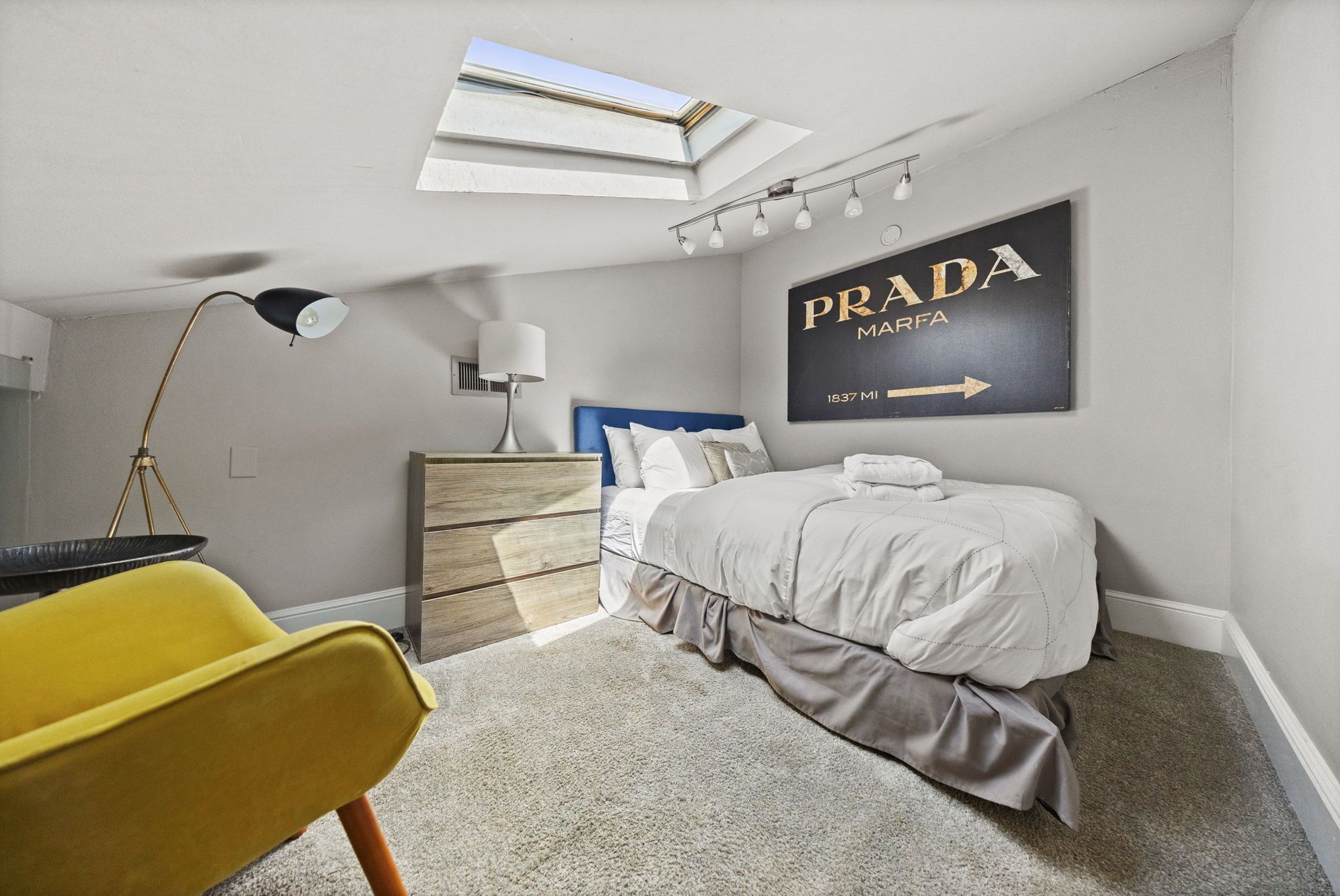 Une chambre à coucher avec un lit, un bureau, une chaise, une lampe, une commode et un tableau accroché au mur.